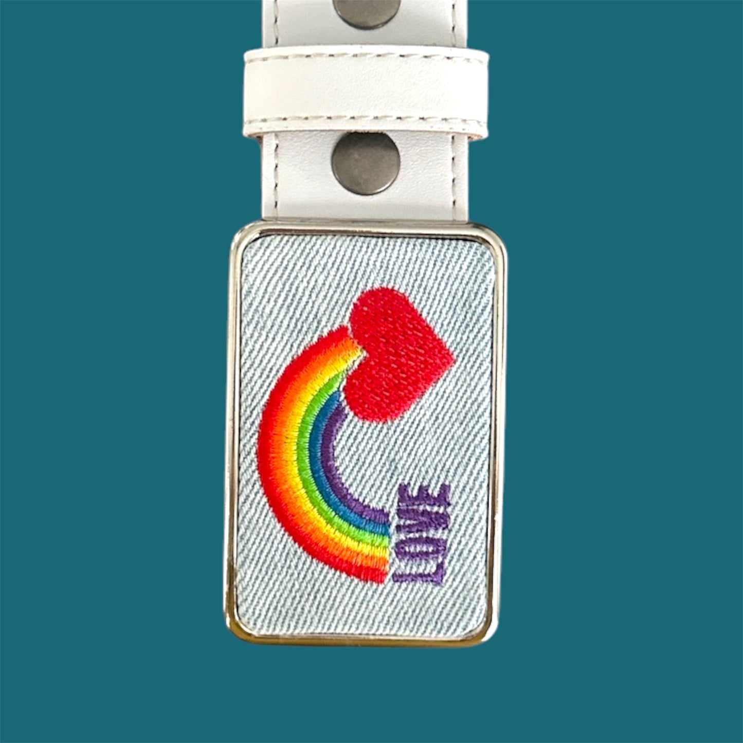 Rectangular embroidered belt buckle with rainbow and heart design on a white strap against a teal background