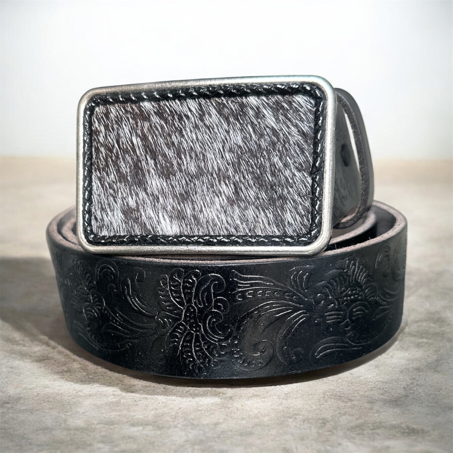 Black leather belt with intricate designs and a metallic hair on hide buckle on a textured surface.