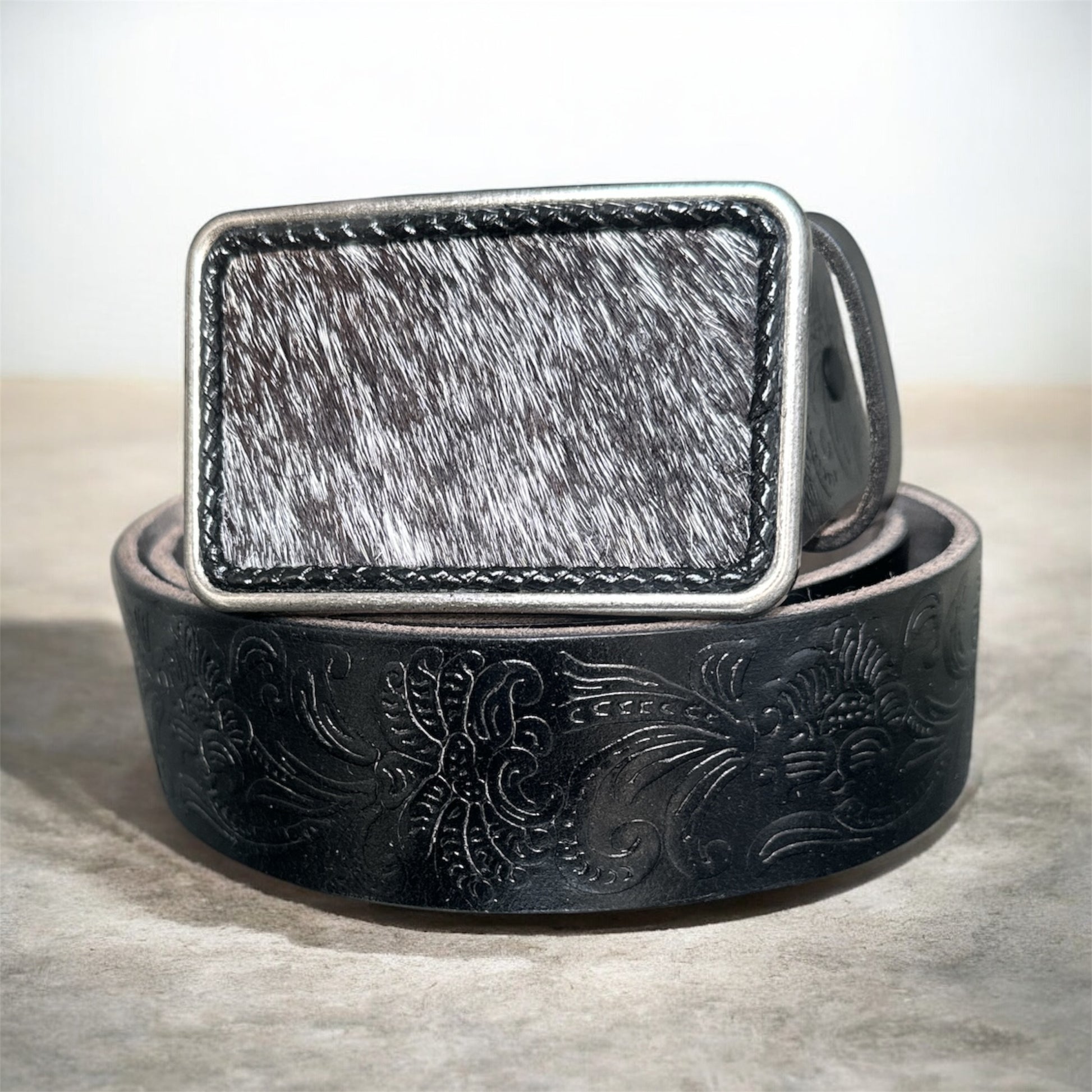 Black leather belt with intricate designs and a metallic hair on hide buckle on a textured surface.
